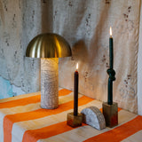 Two gold metal cube candle holders with candles on top and a half-circled clay piece, set on a table beside a lamp