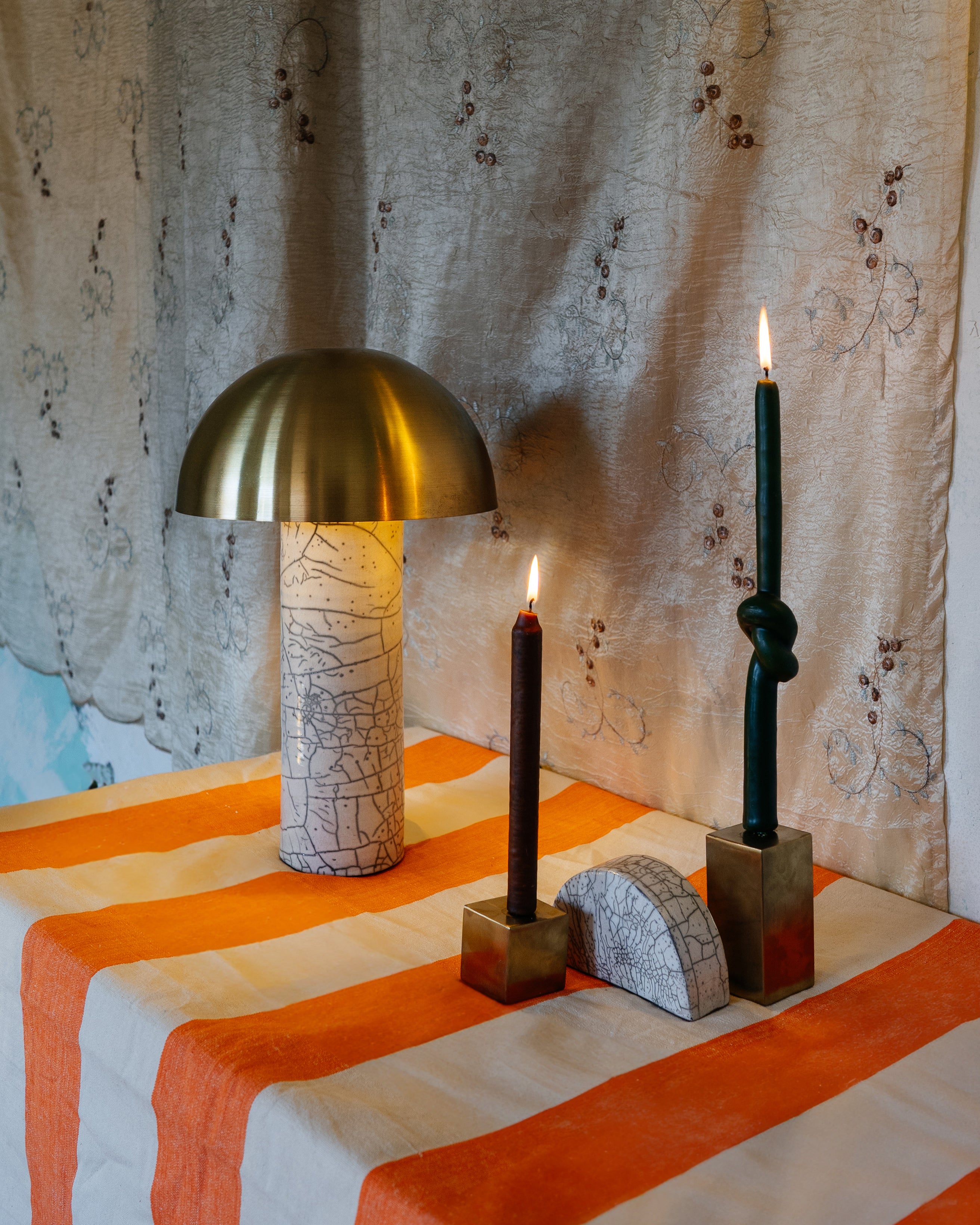 Two gold metal cube candle holders with candles on top and a half-circled clay piece, set on a table beside a lamp