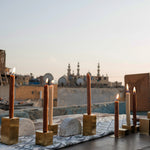 Two gold metal cube candle holders with candles on top, alongside a half-circled clay piece, featured in a photoshoot on a rooftop in Egypt.